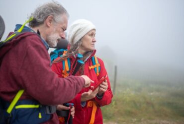 Dois caminhantes com mochilas consultam um telemóvel numa paisagem rural com nevoeiro.