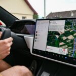 Condutor segura o volante de um Tesla enquanto o ecrã central mostra um percurso de navegação.