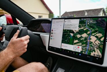 Condutor segura o volante de um Tesla enquanto o ecrã central mostra um percurso de navegação.
