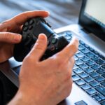 Pessoa segura um controle de videogame diante de um laptop aberto sobre o teclado.