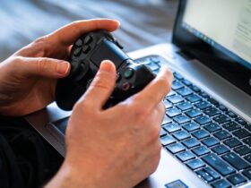 Pessoa segura um controle de videogame diante de um laptop aberto sobre o teclado.