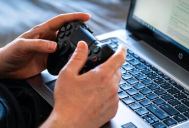 Pessoa segura um controle de videogame diante de um laptop aberto sobre o teclado.