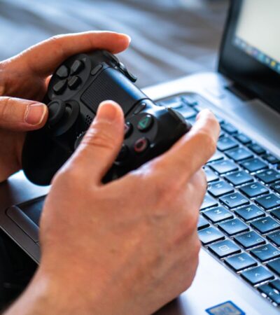Pessoa segura um controle de videogame diante de um laptop aberto sobre o teclado.
