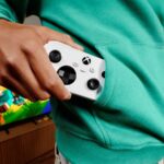 Pessoa guarda um comando Xbox Nano branco no bolso de uma camisola verde, com um televisor ao fundo.