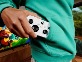 Pessoa guarda um comando Xbox Nano branco no bolso de uma camisola verde, com um televisor ao fundo.