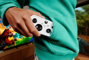Pessoa guarda um comando Xbox Nano branco no bolso de uma camisola verde, com um televisor ao fundo.
