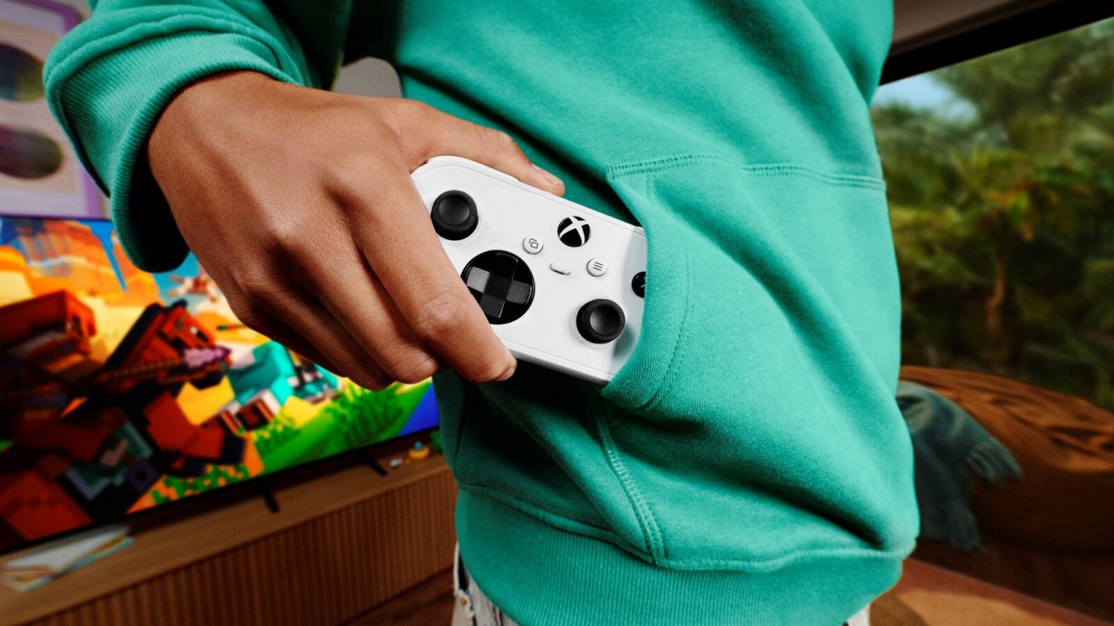Pessoa guarda um comando Xbox Nano branco no bolso de uma camisola verde, com um televisor ao fundo.